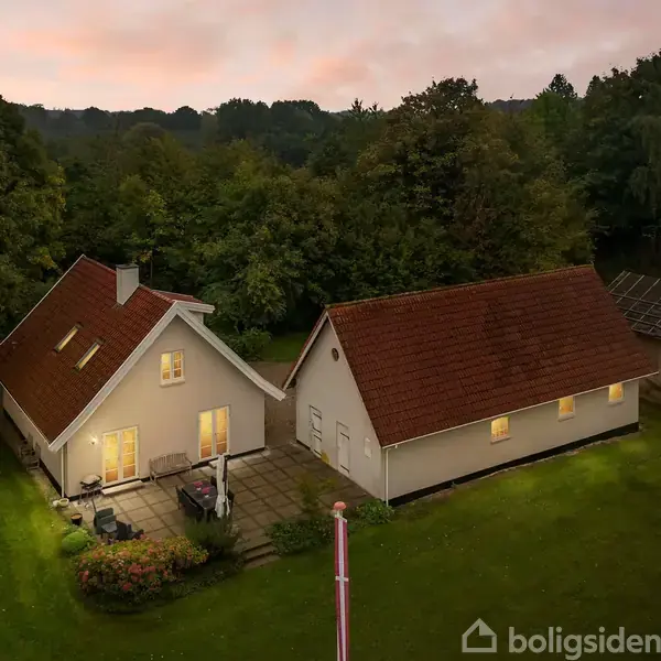 To huse med oplyste vinduer står i en frodig have, omgivet af tætte træer i skumringen. Der er en terrasse med møbler i forgrunden.