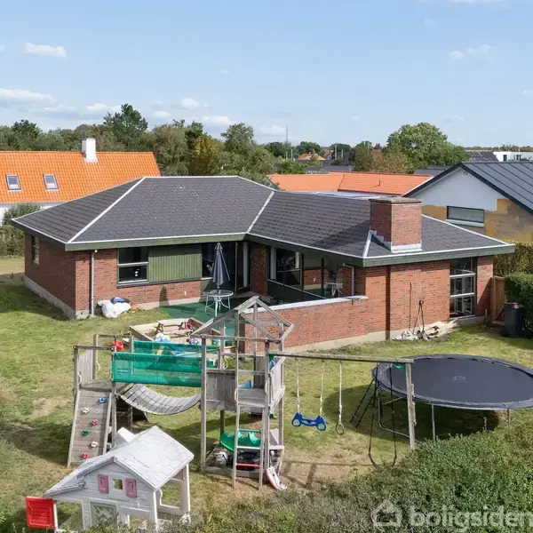 Rød murstensvilla med grått tag i en have med legetårn og trampolin. Omgivet af hække og naboers huse med farverige tage under en blå himmel.