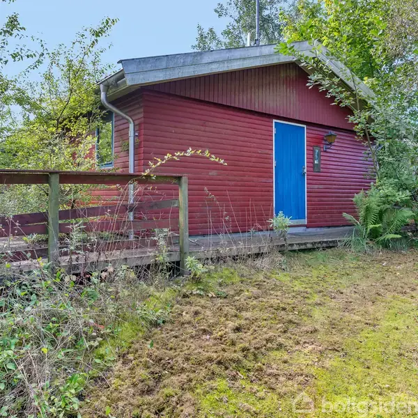 Rødt træhus med blå dør står blandt træer og buske. Foran huset er der en lille veranda omgivet af vildtvoksende natur. Ingen tekst er synlig på billedet.