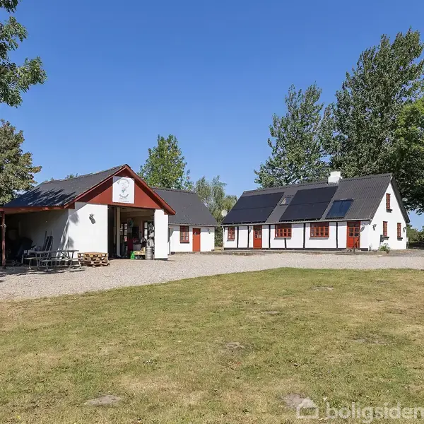 Hvidt bondehus med rødt tag og carport til venstre, omgivet af græsplæne og grønne træer under en klar blå himmel. En bil holder parkeret under carporten.