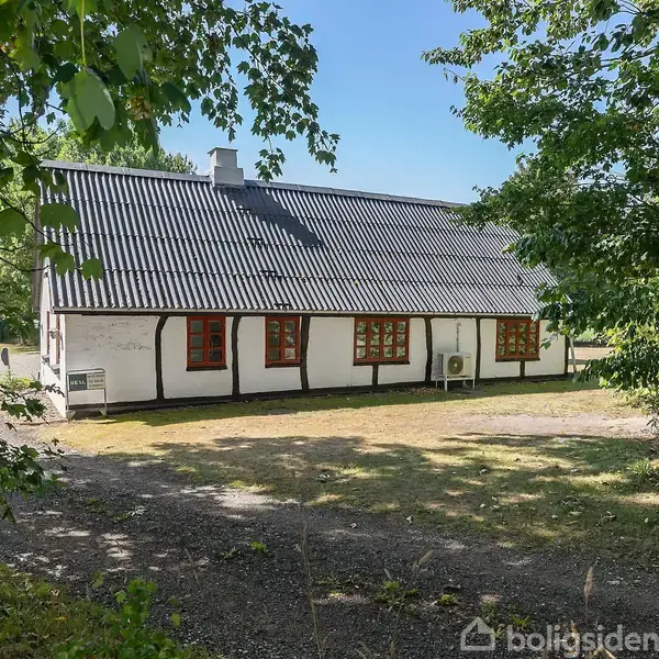 Et bindingsværkshus med sort tag og små vinduer står omgivet af frodig grøn bevoksning på en lysning. Gruset jord omgiver huset hvor sollys filtrerer gennem træernes grene.