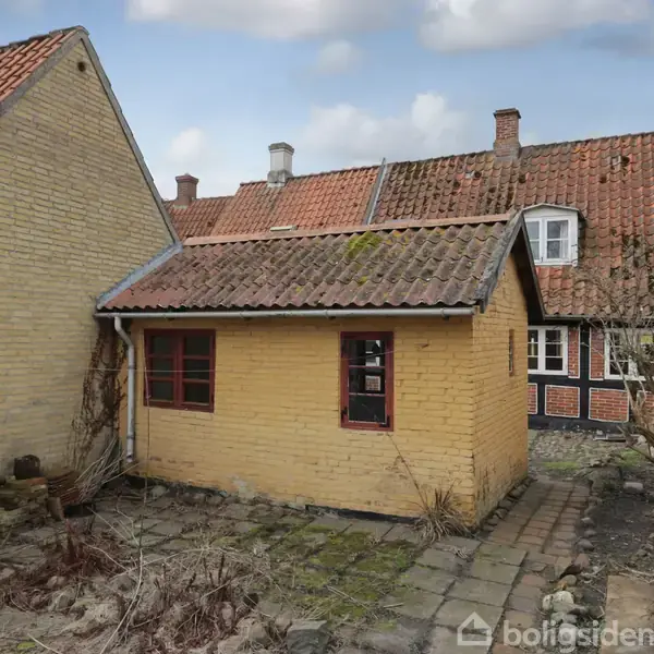 Et lille gult murstenshus med rødt tegltag står på en brolagt gårdsplads omgivet af andre ældre huse. Der er tørre planter langs huset, og nogle vinduer er synlige.