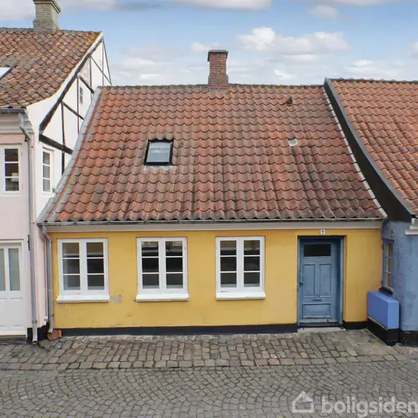 Gult hus med tre hvide vinduer og blå dør står i række af farverige huse. Brostensfortov foran og skyer på himlen.