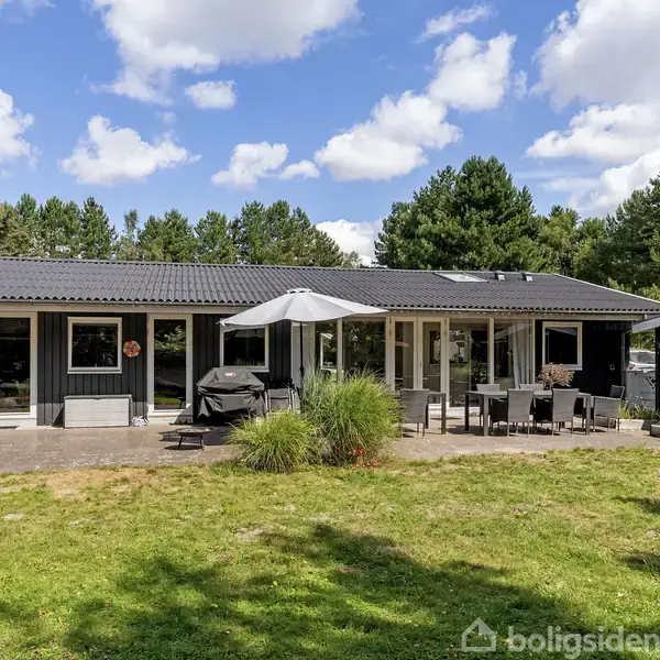 Sort-malet hus med terrasse møbleret med parasol og havemøbler, i en grøn have omgivet af træer. Skyfri himmel med spredte skyer danner baggrund.