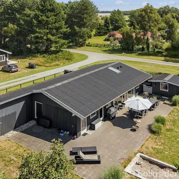 Et sortmalet træhus med fladt tag står i en grøn have. Møbler og parasoller ses på terrassen foran huset. Omkring er der træer og en lille vej.