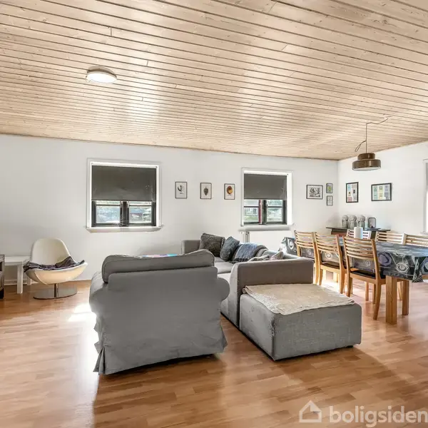 Sofa står i en lys stue med lakeret trægulv og træloft, ved siden af et spisebord med stole. To vinduer har rullegardiner, og der hænger billeder på væggen.