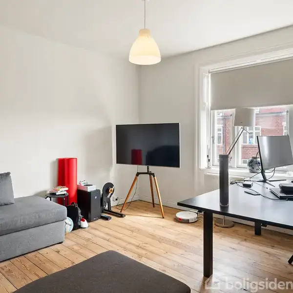 En grå sofa står i et rum ved siden af et sort TV på stativ. Der er et skrivebord med computer og skærm ved et vindue. Trægulve og lyse vægge.
