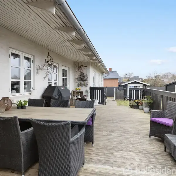 Udendørs terrasse med et bord og stole i flet, omgivet af høje træhegn. Blomsterkrukker og en grill står tæt på husets hvide facade. Himmel blå og klar.