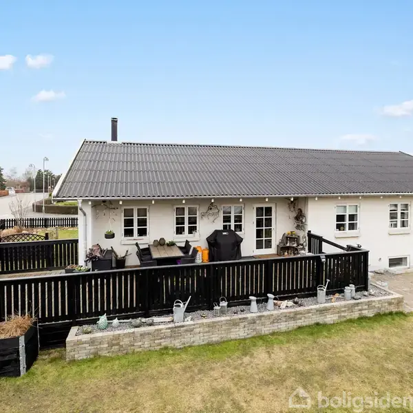 Hus med sort tag og terrasse. Terrasse omgivet af sort stakit og dekoreret med planter. Husets omgivelser inkluderer græsplæne og vej i et forstadsområde.