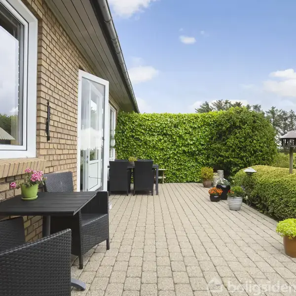 Terrasse med flisebelægning. To sorte borde med stole står opstillet. Grønne planter i krukker og buske omkranser terrassen. Husmur af gule mursten til venstre og grøn bevoksning i baggrunden.