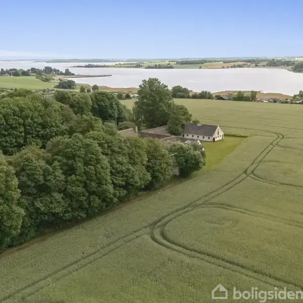En gård med stråtækte bygninger omgivet af frodige træer står stille i et åbent landskab med marker og grønne områder i baggrunden.