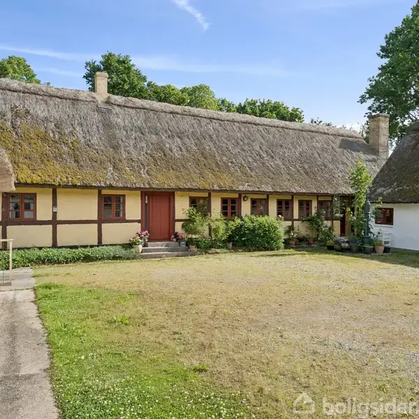 En hvid bygning står blandt træer ved en mark. Bygningen ligger i et landligt område med udsigt over en sø i baggrunden.