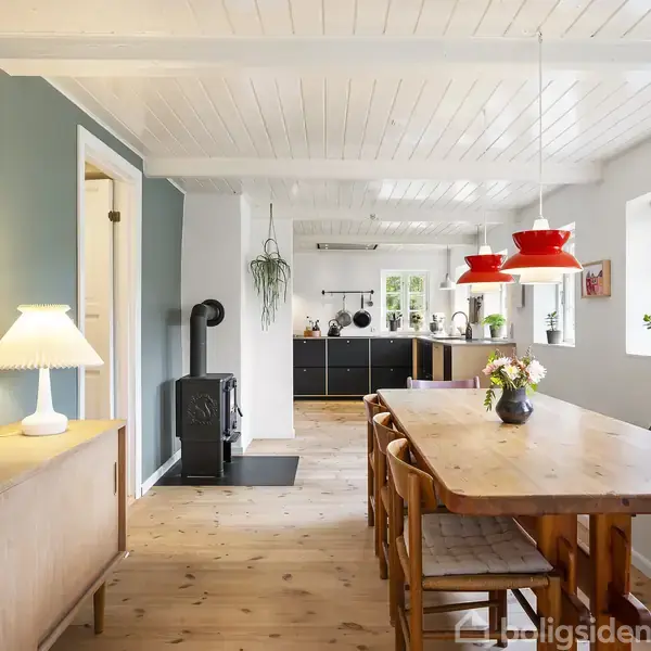Træbord med vase og stole står i en lys spisestue. Røde lamper hænger over bordet, omgivet af lyse trævægge og hvide vinduer. En plakat med teksten “SJÆLLAND FREDERIKSDAL HAVEN ULSLEV” hænger på væggen.