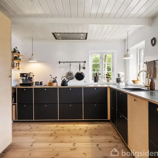 Et moderne køkken med sorte skabe og lyse træoverflader, hvor køkkenredskaber hænger ved en væg. Rummet har hvide vægge og lofter, trægulv og store vinduer, der giver naturligt lys.
