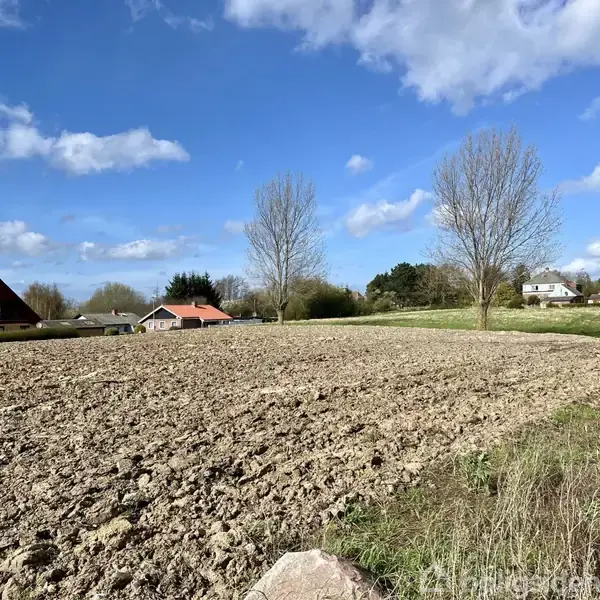 En pløjet mark ligger stille i et landskab med et par træer og spredte huse i baggrunden under en blå himmel med skyer.