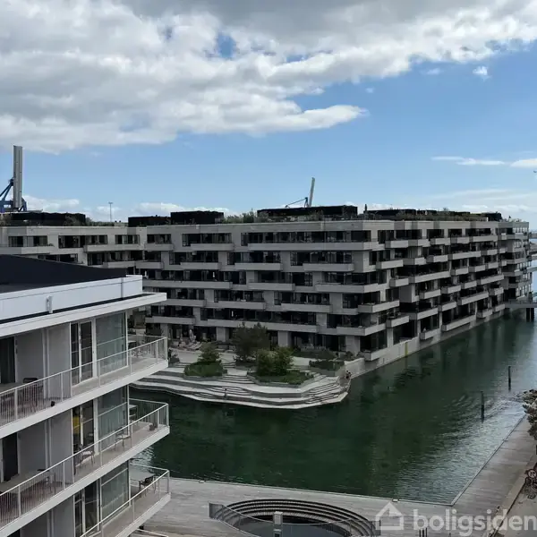 Lejlighedskomplekser ligger ved en kanal i en havneby. De moderne bygninger spejler sig i vandet, mens en bro ses i baggrunden under en delvist skyet himmel.
