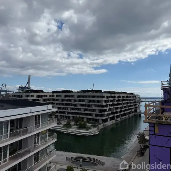 Moderne lejlighedskompleks strækker sig langs en vandkanal, omgivet af flere etager og byggestilladser. I baggrunden er kraner synlige mod en delvist overskyet himmel.
