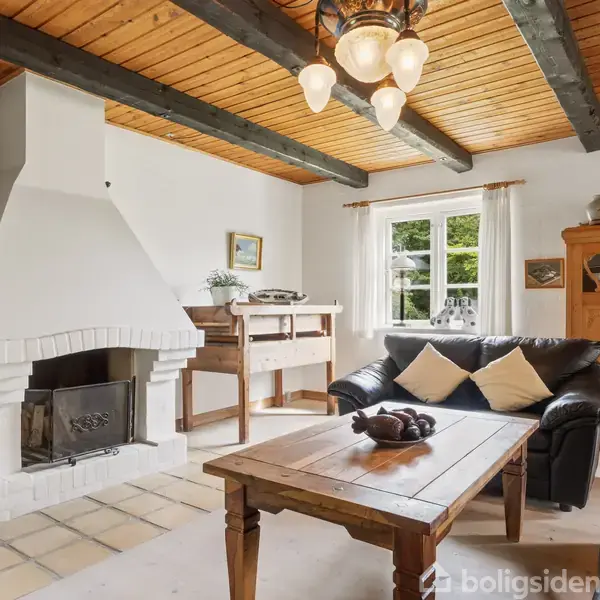 En hyggestue med en hvid pejs i hjørnet, sofaer omkring et træbord, og et skab med dekorative genstande ved siden af et vindue, der slipper naturligt lys ind.