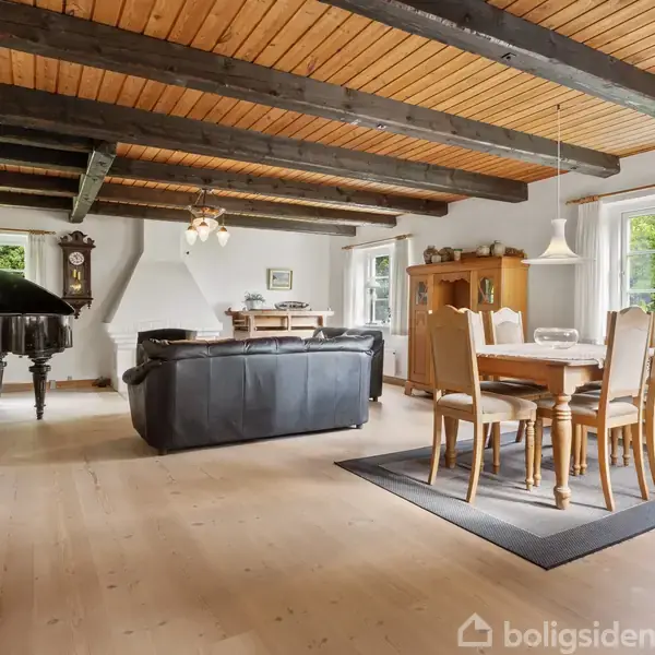 Et flygel står i en stue og giver en elegant atmosfære; omkring ses spisebord, sofa, lyse lofter med mørke bjælker og store vinduer, hvilket skaber et lyst og indbydende rum.