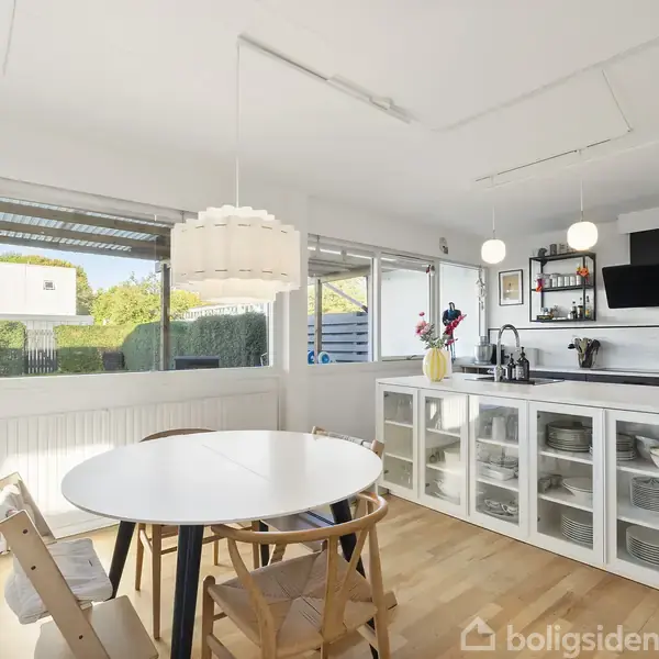 Et lyst køkken med et rundt spisebord og tre stole. Køkkenø med indbyggede opbevaringspladser. Rummelige vinduer giver udsigt til en grøn have.