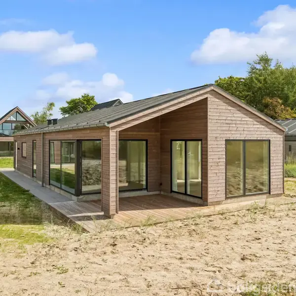 Træhus står stille på en sandet grund med store vinduer. Ved siden af er et andet hus med trekantede gavle. Grønne træer ses i baggrunden under en blå himmel.
