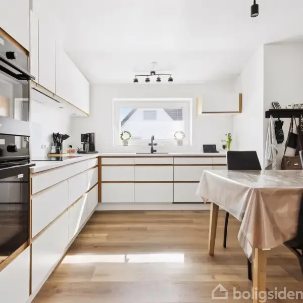 Et moderne køkken med hvide skabe og integrerede ovne. En bordplade viser en kaffemaskine; et bord med dug og stole står centralt. Et vindue slipper naturligt lys ind.