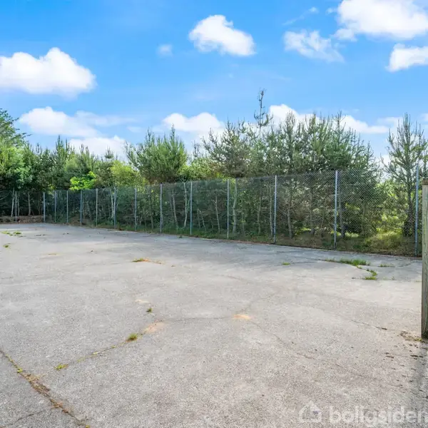 Betonplads med to fodboldmål, uden aktivitet, omgivet af hegn og træer, under en blå himmel med skyer.