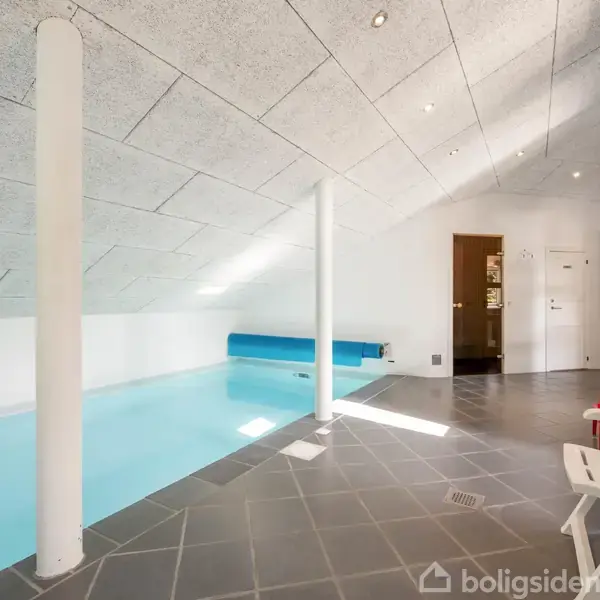 Indendørs swimmingpool med blå vand under en skrånende loft. To hvide søjler står ved siden af poolen. Omkring er der grå fliser, to hvide stole og en rød strandstol.