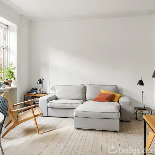 Grå sofa står i en lys stue ved vindue. Tæt på ses en træstol og et bord med lampe. Væggen er hvid, og der er farverige puder på sofaen.