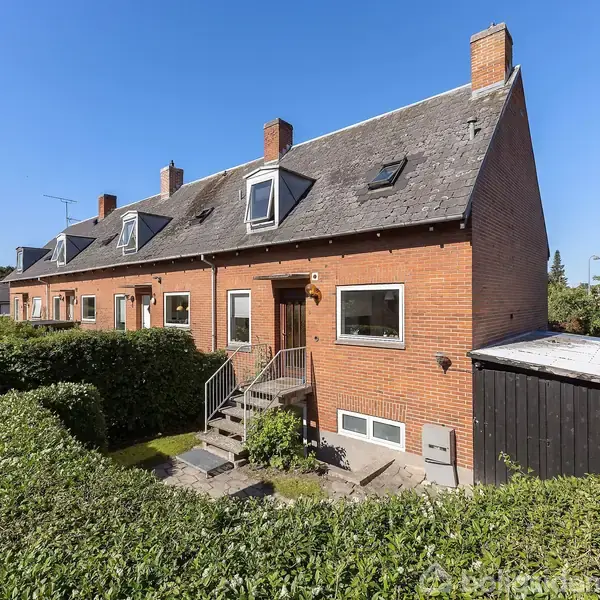 Rækkehus i mursten med skråt tag og kvistvinduer, omgivet af en hæk i et boligområde. Trappe fører op til hoveddøren. Klar blå himmel indikerer solrigt vejr.