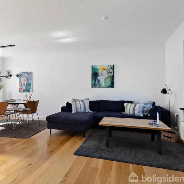 Sort hjørnesofa står mod en hvid væg, dekoreret med kunstmalerier. Foran sofaen er der et sofabord. I baggrunden er et spisebord med stole, og et stort vindue skaber lysindfald.