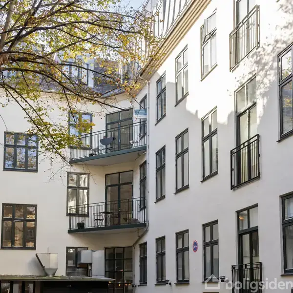 En hvid bygning med mange vinduer, balkoner og en stor træ i forgrunden. Bygningen ser ud til at være i et boligområde.