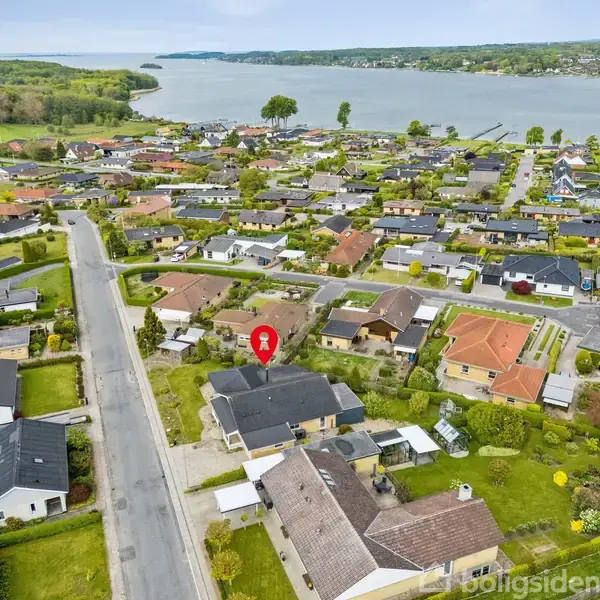 Et beboelseshus markeret med en rød pin. Det befinder sig i et boligområde med mange huse og grønne haver. Baggrunden viser en sø og skovklædte områder i horisonten.