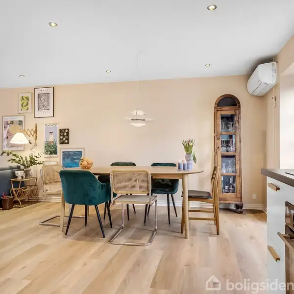 Spisebord står centralt med forskellige stole omkring. En vase med blomster er på bordet. Rummet har lyse trægulve, vægkunst og et skab samt et opholdsområde i baggrunden.