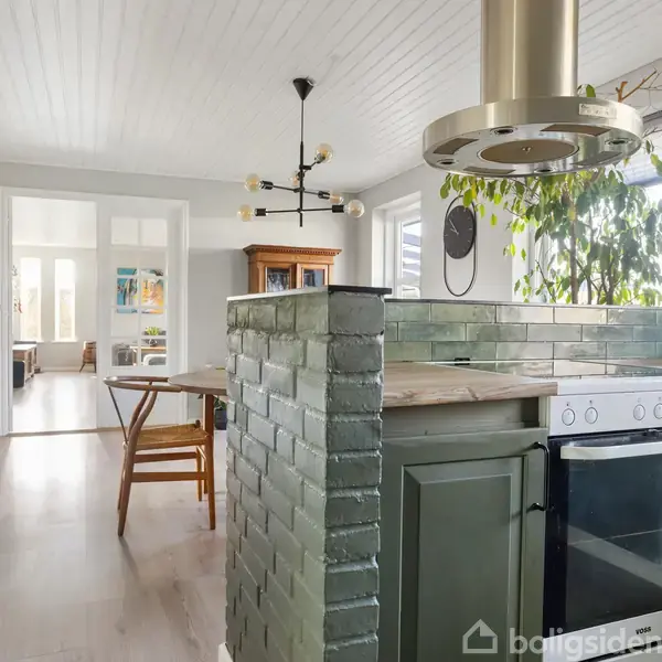 Køkken med grønne fliser og moderne emhætte ovenpå indbygningsovn. Rundt spisebord med træstol. Indrammede kunstværker på væggen. Store vinduer og grønne planter ved siden.