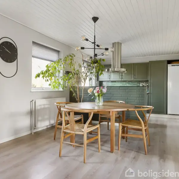 Rundt spisebord med fire stole står i køkken-alrum. En stor plante og et ur hænger ved vinduet. Baggrunden viser et grønt køkken og en hvid køleskab. Vase med blomster på bordet.
