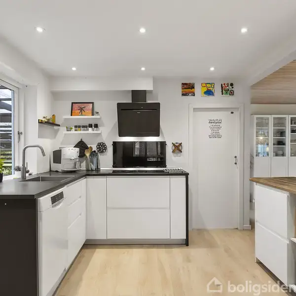 Moderne køkken med hvide skabe og sort bordplade, indeholder indbygget ovn og komfur. Hylder med dekoration hænger over. Dør med tekst: "Jespers madbod: ... sagt kan ikke bruges mod mig".