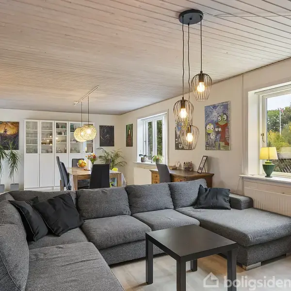 Grå sofa står i stue med lys træloft og vinduer mod haven. Lamper hænger over sofabord, og spisebord ses i spisestue med malerier og planter på vægge.