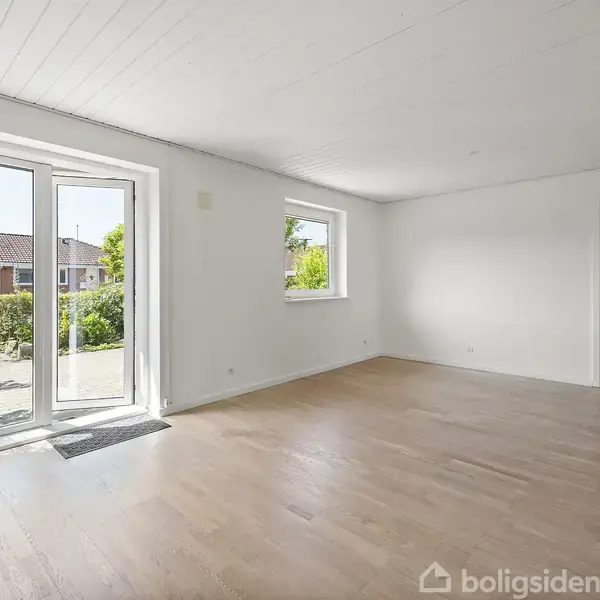 Stue med trægulv og hvidmalede vægge, store glasdøre åbner mod en terrasse med udsigt til en have. Små vinduer giver ekstra lys.