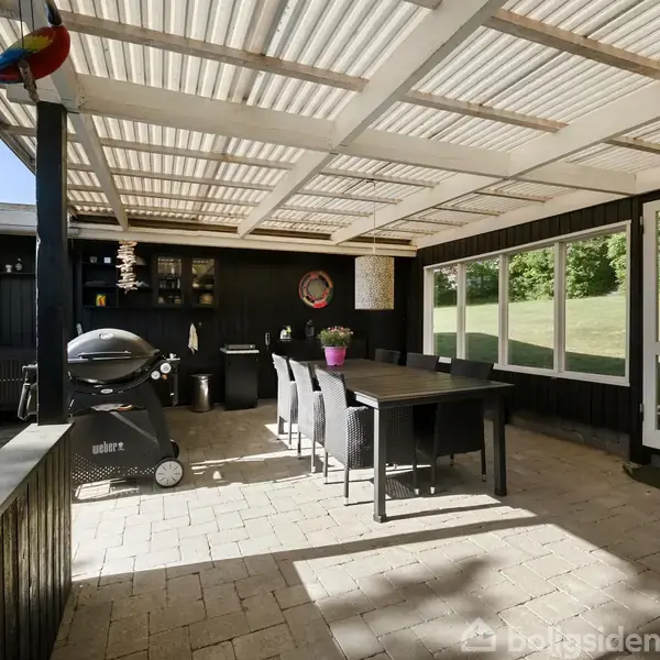 Terrasse med sort spisebord og seks stole under halvtag. En Weber grill står ved siden af bordet. Bagved er en murstensvæg med vinduer og dør, græsplæne udenfor.