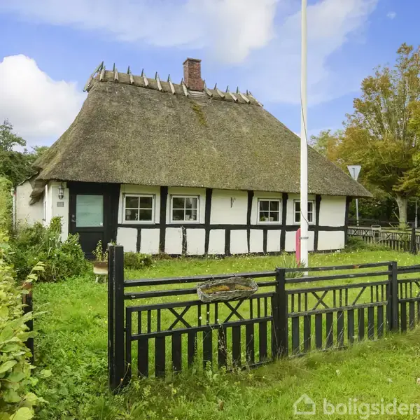 Bindingsværkshus med stråtag; huset står stationært i en grøn have med et sort trægærde foran. Omgivet af træer og en lys himmel.