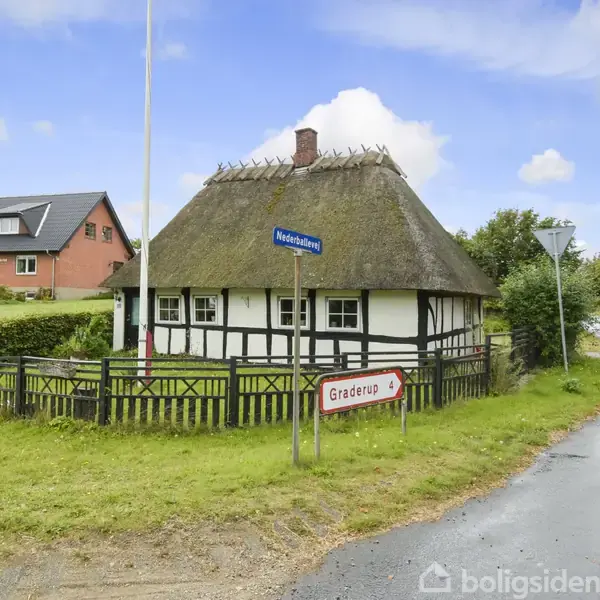 Et bindingsværkshus med stråtag står ved en vej. Et skilt foran viser "Graderup". En grøn have omgiver huset, og en anden bygning kan ses i baggrunden.