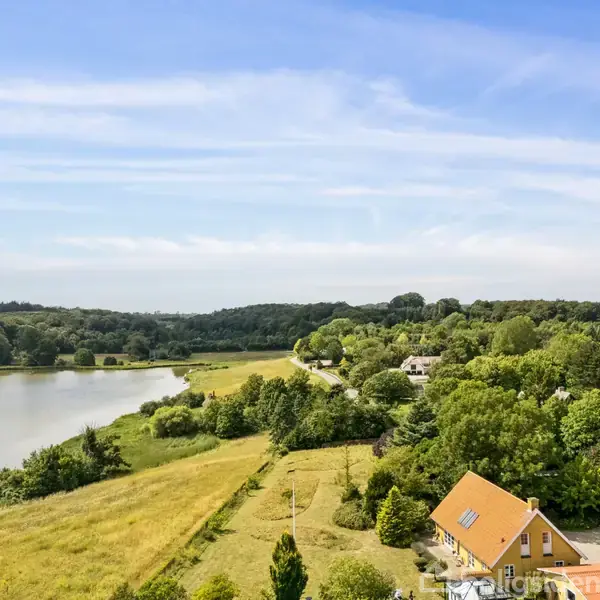 Et hus med rødt tag ligger blandt træer i en frodig landskabskontekst ved siden af en stor sø. Skovklædte områder og græsmarker omringer søen under en blå himmel.