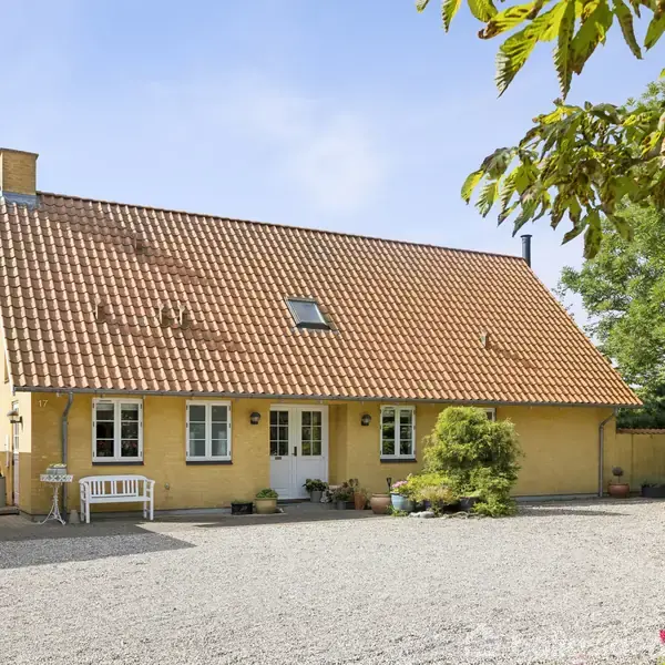 Gult hus med rødt tegltag står på en grusplads. En hvid bænk og planter pynter foran huset. En gul garage med tallet "17" ses til venstre, omgivet af grønne træer.