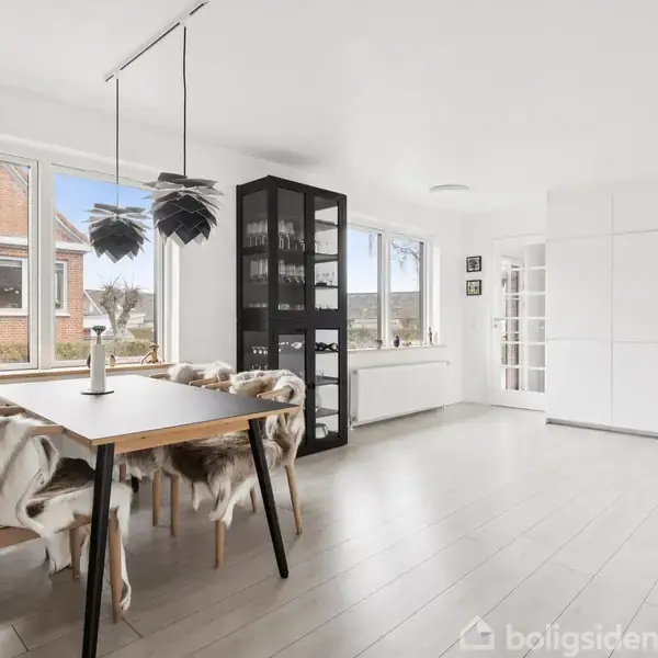 Spisebord med sorte stole dækket af pels, står i en lys spisestue med hvide vægge og gulve. Store vinduer giver udsigt til et hus udendørs. En sort vitrineskab står i hjørnet.