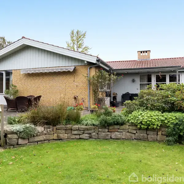 Et murstenshus med terrasse, omgivet af grønne planter og en græsplæne. Træmøbler står på terrassen. Baggrunden viser en blå himmel og nogle træer.