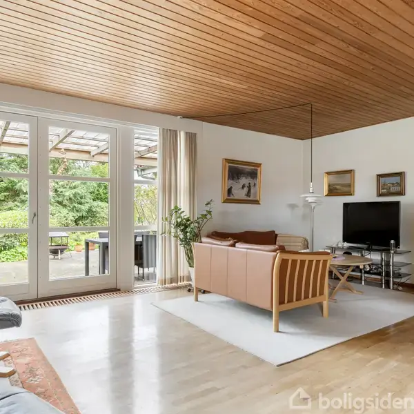 En stue med en sofa og fladskærms-TV, handlingen er stilstand. Konteksten er et lyst rum med trægulv, træloft, store vinduer med udsigt til en grøn have og flere malerier på væggen.