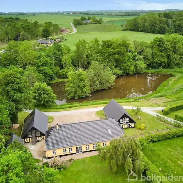 Luftfoto af en gård med tre bygninger omgivet af grønne marker og skov. En lille sø er placeret i nærheden af træerne, og en snoet vej fører til ejendommen.