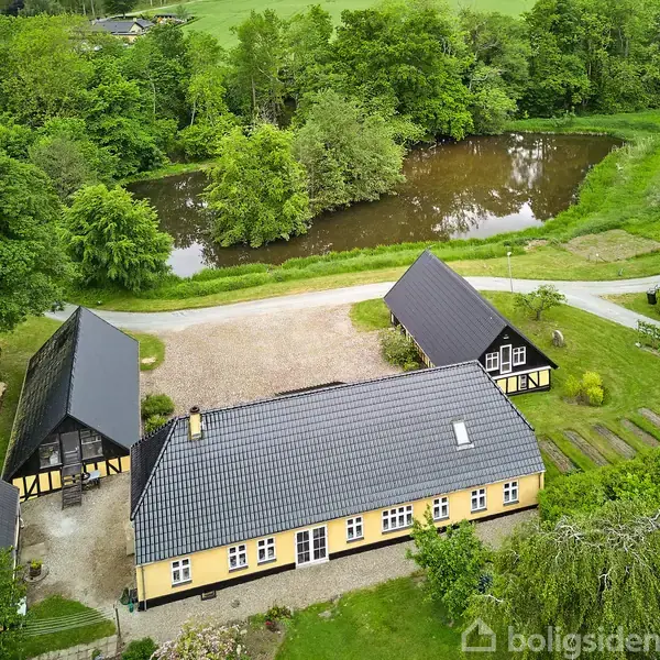 Gult bindingsværkshus med mørkt tag står stille omgivet af frodig have og græsarealer. En dam og grønne træer danner en landskabelig baggrund i naturskønne omgivelser.