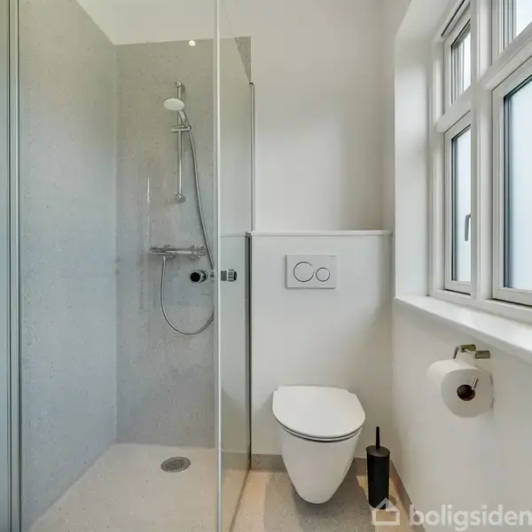 Brusekabine med bruser i gang, ved siden af væghængt toilet, omgivende af lyse vægge. Vindue til højre med naturligt lys. Toiletrulleholder og toiletkumme er synlige.